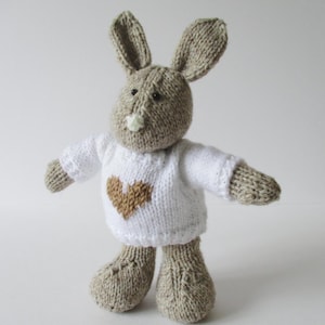 Puede incluir: Un conejo de peluche tejido a mano con hilo marrón claro, orejas largas y ojos de cuentas negras. El conejo lleva un suéter blanco tejido con un diseño de corazón marrón en el pecho. El juguete tiene una nariz clara y pies texturizados.
