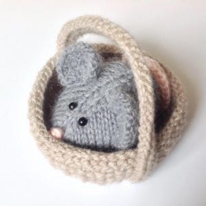 Op de afbeelding: Een kleine, handgebreide grijze muis met zwarte kraaloogjes en een piepklein roze neusje, genesteld in een rond, beige gehaakt mandje met een hengsel. De muis is gemaakt van zacht garen en het mandje heeft een gestructureerd, dik steekpatroon.