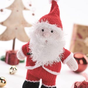 Pode incluir: Uma figura de Papai Noel de malha, em um traje vermelho com barba e chapéu brancos. A boneca do Papai Noel tem botas pretas e acabamentos brancos. Decorações de Natal, incluindo uma árvore e ornamentos, no fundo.