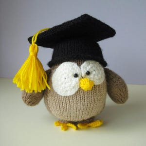 May include: A round, brown knitted owl toy featuring large white eye patches, a small yellow beak, and yellow feet. The owl wears a black knitted graduation cap with a bright yellow tassel hanging from the side, crafted from yarn.