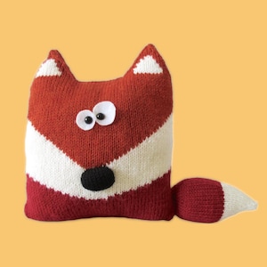 May include: A knitted fox-shaped pillow in red, white, and black. The pillow features a red body with a white stripe, white-tipped ears, and a tail. It has googly eyes and a black nose. The pillow is a decorative item.