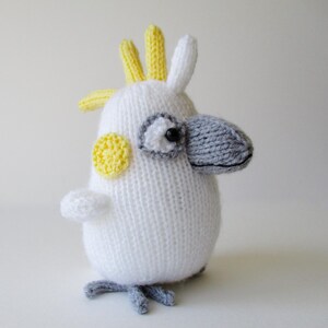 May include: A hand-knitted cockatoo plush toy featuring a white body, a prominent grey beak, and a yellow crest. The bird has a small yellow circular patch on its cheek, a black eye, and grey feet. The texture is consistent with yarn knitting.