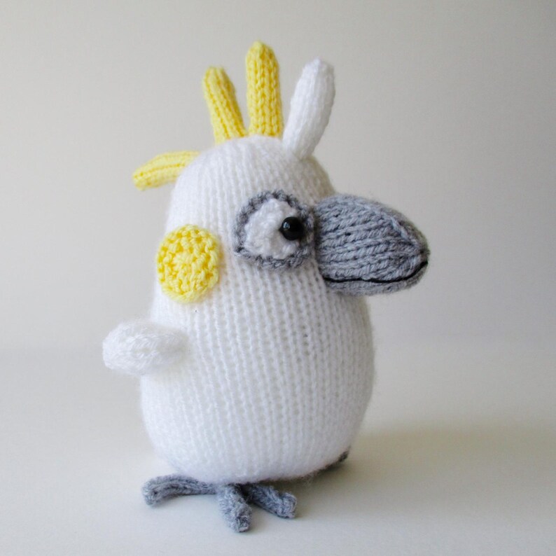 May include: A hand-knitted cockatoo plush toy featuring a white body, a prominent grey beak, and a yellow crest. The bird has a small yellow circular patch on its cheek, a black eye, and grey feet. The texture is consistent with yarn knitting.