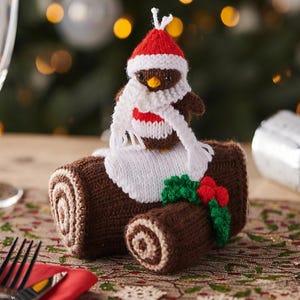 May include: A knitted brown yule log cake decoration featuring a small brown bird wearing a red and white hat and a white scarf. The log has a white knitted patch on top, green holly leaves, and red berries attached to the side of the brown structure.