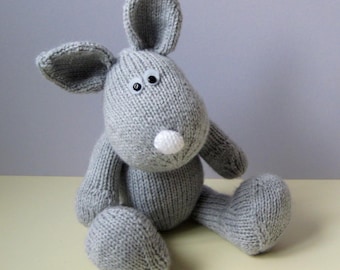 May include: A grey knitted stuffed animal shaped like a rabbit with two long ears, a round white nose, and two small black plastic eyes. The toy is constructed from a textured knit fabric and is seated in an upright position with legs extended.