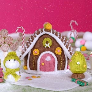 May include: A knitted Christmas scene featuring a brown gingerbread house with white trim, a pink door, and yellow flower accents. A snowman with a green hat and scarf, and a green Christmas tree are also present. Candy canes and snowballs complete the festive display.