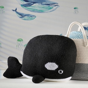 May include: A black and white knitted orca whale plush toy with a white belly and eye details. The whale is positioned in front of a basket and a wall with whale and jellyfish illustrations.