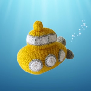 May include: A yellow knitted submarine toy featuring a white conning tower with grey trim, three circular white portholes with grey borders along the side, and a grey propeller at the rear. The soft textile object has a rounded, plush construction.