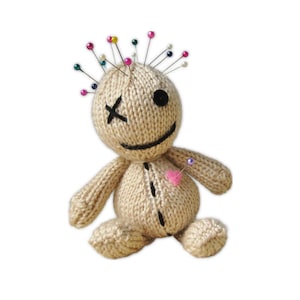 May include: A beige knitted voodoo doll with a black cross for an eye and a black button eye. The doll has a stitched smile and a pink heart pin in its chest. Various coloured pins are inserted into its head.