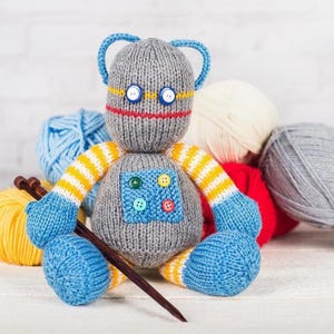May include: A handmade grey knitted robot doll with blue details, yellow and white striped arms, and a blue square patch with colourful buttons. The robot has blue ears, button eyes, and a red stitched mouth. Knitting needles and yarn balls are in the background.