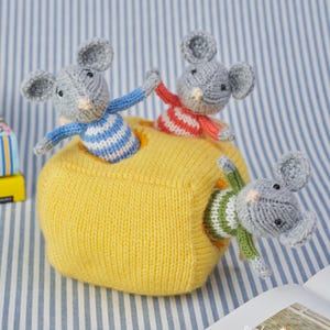 May include: A handmade knitted toy featuring three gray mice emerging from a yellow knitted ball. The mice wear blue and white, red and white, and green and white striped shirts. The background is a blue and white striped fabric.