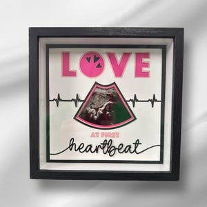 May include: A black picture frame with a white background and the words "LOVE AT FIRST HEARTBEAT" in pink and black lettering. A black and white ultrasound image is inside the frame.