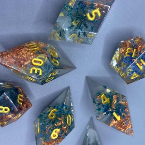 Peut inclure: Un ensemble de sept dés polyédriques en résine transparente. Les dés présentent des chiffres dorés et des inclusions bleues et cuivrées. Ces dés sont conçus pour les jeux de rôle sur table.