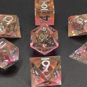 May include: Set of seven polyhedral dice with a pink and gold color scheme. The dice are transparent with gold glitter and pink hues. Each die has silver numbering. The dice are for tabletop games.