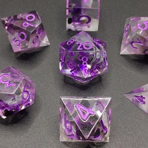 May include: Set of seven clear, purple-numbered polyhedral dice for tabletop games. The dice have a marbled purple and white interior, with the numbers painted in a vibrant purple. The dice include a D4, D6, D8, D10, D12, D20, and a percentile die.