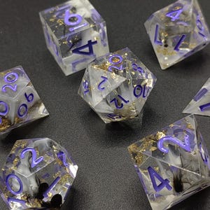 Peut inclure: Un ensemble de sept dés polyédriques transparents, parfaits pour les jeux de rôle. Les dés présentent un motif tourbillonnant blanc et noir, des chiffres violets et des accents dorés. L'ensemble comprend un d4, d6, d8, d10, d12, d20 et un dé de pourcentage.