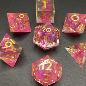 Peut inclure: Ensemble de sept dés polyédriques. Chaque dé est transparent avec des inclusions roses et violettes et des chiffres dorés. Les dés sont conçus pour les jeux de rôle sur table.