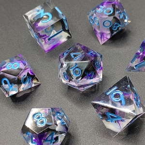Peut inclure: Ensemble de sept dés polyédriques pour jeux de rôle. Les dés transparents présentent un mélange tourbillonnant de violet, noir et blanc. Les chiffres sont d'un bleu vif. Les dés sont posés sur une surface gris foncé.