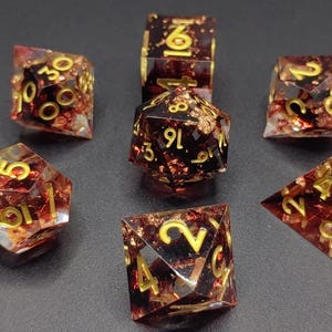 May include: Set of seven polyhedral dice for tabletop games. The dice are transparent with a reddish-brown hue, gold numbers, and gold leaf inclusions. The dice are arranged on a dark surface.