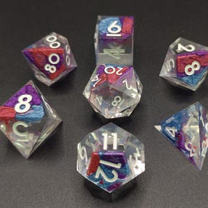Peut inclure: Un ensemble de sept dés en résine transparente avec des accents rouges, bleus et violets incrustés. Chaque dé comporte des chiffres blancs. Les dés sont conçus pour les jeux de rôle sur table et sont présentés sur un fond sombre.
