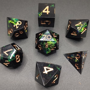 May include: A set of seven black polyhedral dice with gold numbering and iridescent green and blue inclusions. The dice are arranged on a gray surface, showcasing their various shapes, including a d20, d12, d10, d8, d6, and d4.