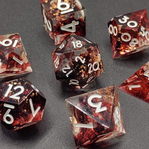 May include: Set of seven polyhedral dice. The dice are clear with a red and gold flecked interior. Each die has white numbering. The dice are arranged on a dark surface.