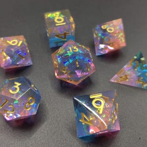 Pode incluir: Um conjunto de sete dados poliédricos em várias formas, incluindo um d4, d6, d8, d10, d12, d20 e um dado percentual. Os dados são transparentes com um efeito iridescente colorido e numeração dourada. O esquema de cores inclui rosa, azul e dourado.