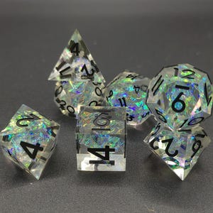 Könnte beinhalten: Set aus sieben durchsichtigen, irisierenden Würfeln mit schwarzen Zahlen. Die Würfel haben verschiedene Formen, darunter eine vierseitige Pyramide, ein sechsseitiger Würfel, ein achtseitiger Würfel, ein zehnseitiger Würfel, ein zwölfflächiger Würfel und ein zwanzigflächiger Würfel.