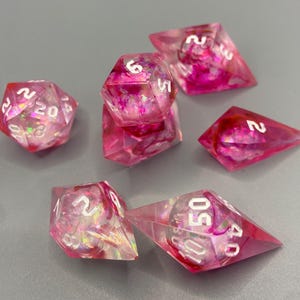 May include: A collection of seven polyhedral dice, ideal for role-playing games. The dice are made from pink and clear resin, with white numbering. They are displayed on a grey surface.