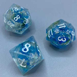 Peut inclure: Trois dés polyédriques avec un motif de paillettes bleues et argentées. Les dés sont transparents avec des chiffres blancs. Les dés sont pour les jeux de société comme Donjons et Dragons.