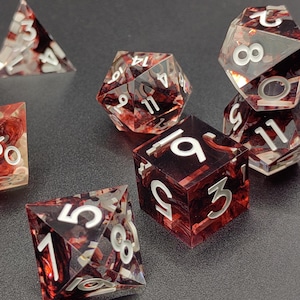 Könnte beinhalten: Ein Set aus sieben polyedrischen Würfeln für Tabletop-Spiele. Die Würfel sind transparent mit einem rot-schwarzen Marmoreffekt und weißen Zahlen. Enthält einen D4, D6, D8, D10, D12, D20 und einen Prozentwürfel.