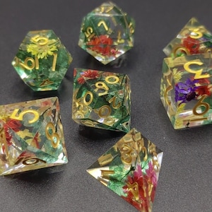 May include: Set of seven polyhedral dice with embedded flowers and gold numbering. The dice are clear with green, yellow, and red floral inclusions. The dice are designed for tabletop games.