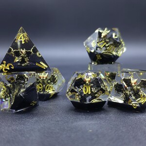 Puede incluir: Un juego de dados poliédricos de resina transparente con bases negras, cada uno con una criatura alada en miniatura de color dorado y ojos rojos. Los números están grabados en oro sobre diversas formas geométricas.