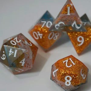 Op de afbeelding: Een verzameling handgemaakte veelvlakkige dobbelstenen in verschillende vormen. De dobbelstenen hebben glinsterend oranje en heldere hars met witte cijfers. De set bevat een D4, D6, D8, D10, D12, D20 en een D100.