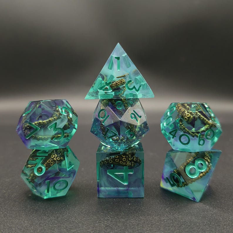 May include: A set of seven polyhedral dice made of translucent teal and purple resin. Each die contains a miniature metallic gold octopus tentacle with visible suckers embedded inside. The numbers are engraved in a matching teal color.