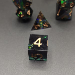 May include: A set of polyhedral dice made from black resin with embedded iridescent green and blue flakes. The numbers are engraved in a metallic gold finish. The set includes a four-sided die, a six-sided die, and other multi-sided shapes.