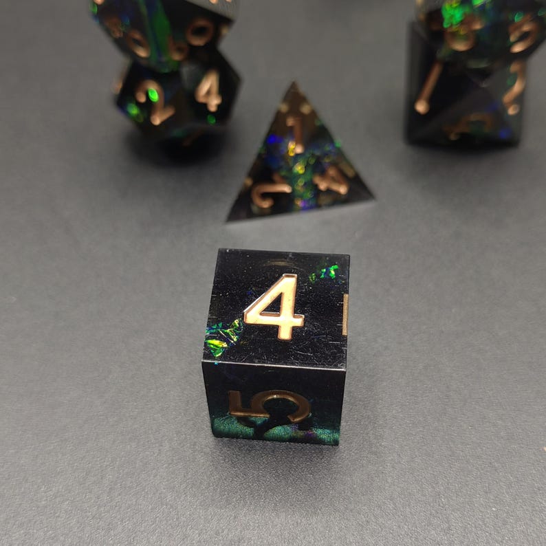 May include: A set of polyhedral dice made from black resin with embedded iridescent green and blue flakes. The numbers are engraved in a metallic gold finish. The set includes a four-sided die, a six-sided die, and other multi-sided shapes.