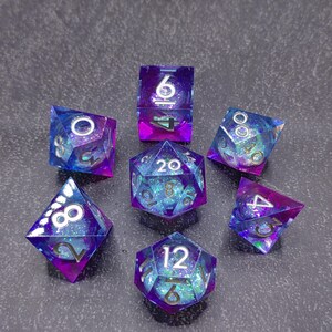 May include: A set of seven polyhedral dice in shades of blue, purple, and green. The dice are transparent with glitter and feature white numbering. The set includes a D4, D6, D8, D10, D12, D20, and a percentile die, perfect for tabletop gaming.