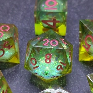 Puede incluir: Primer plano de varios dados poliédricos. Los dados son transparentes con un interior verde y brillante y numeración roja. Los bordes son amarillos. Los dados se utilizan para juegos de rol de mesa.