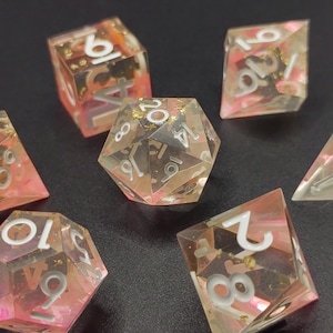 Peut inclure: Un ensemble de dés en résine transparente avec des inclusions roses et dorées. Les dés ont des chiffres blancs et sont de formes géométriques variées, dont un dé à 20 faces. Idéal pour les jeux de rôle.