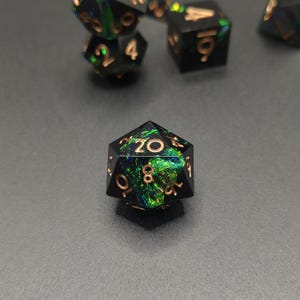May include: A twenty-sided die featuring a black resin base with embedded iridescent green and blue foil flakes. The numbers are engraved in a metallic copper color, with the number twenty prominently displayed at the top center face.