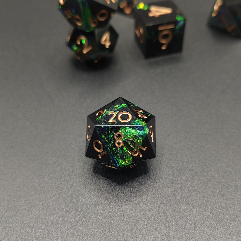May include: A twenty-sided die featuring a black resin base with embedded iridescent green and blue foil flakes. The numbers are engraved in a metallic copper color, with the number twenty prominently displayed at the top center face.