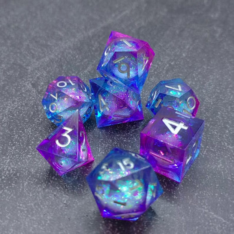 May include: A set of seven polyhedral dice in shades of blue and purple with silver numbering. The dice are made of a clear resin with glitter suspended inside, creating a shimmering effect. The dice are arranged on a gray surface.