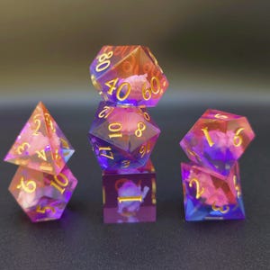 Puede incluir: Juego de seis dados poliédricos en tonos morados, rosas y dorados. Los dados son transparentes con un diseño nublado y floral en su interior. Números dorados visibles en cada dado, con formas variadas: d4, d6, d8, d10, d12 y d20.
