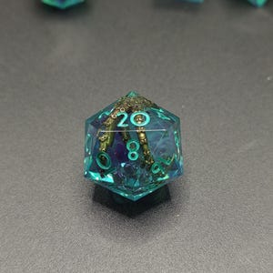 May include: A translucent teal twenty-sided die featuring an embedded metallic bronze tentacle with visible suction cups. The numbers are etched in a bright cyan color, with the number twenty clearly visible at the top of the die face.