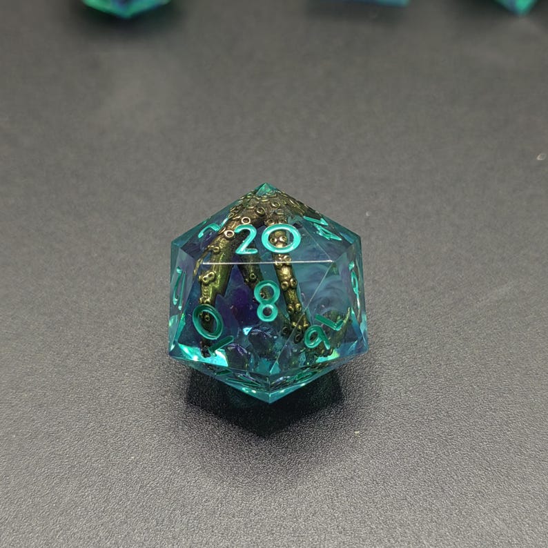 May include: A translucent teal twenty-sided die featuring an embedded metallic bronze tentacle with visible suction cups. The numbers are etched in a bright cyan color, with the number twenty clearly visible at the top of the die face.