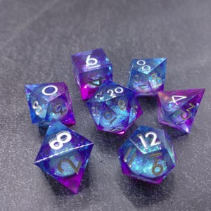 May include: Set of seven polyhedral dice in shades of blue and purple with iridescent glitter. Each die has white numbering. The dice are designed for tabletop role-playing games.