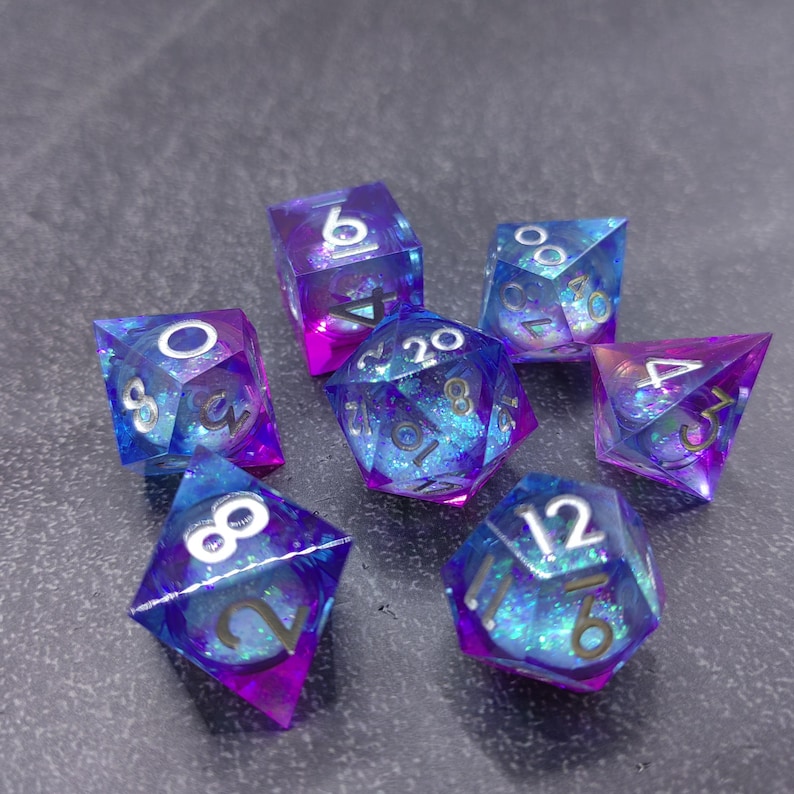 May include: Set of seven polyhedral dice in shades of blue and purple with iridescent glitter. Each die has white numbering. The dice are designed for tabletop role-playing games.