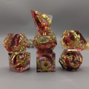 Puede incluir: Juego de siete dados poliédricos rojos y dorados. Los dados transparentes presentan números y detalles dorados. Estos dados están diseñados para juegos de rol de mesa, cada dado tiene un número diferente de lados.
