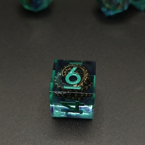 May include: A translucent teal and blue square die featuring an embedded metallic bronze octopus tentacle. The die displays the numbers 4 and 6 in a bright teal font, with the tentacle curling around the numeral 6 inside the resin.